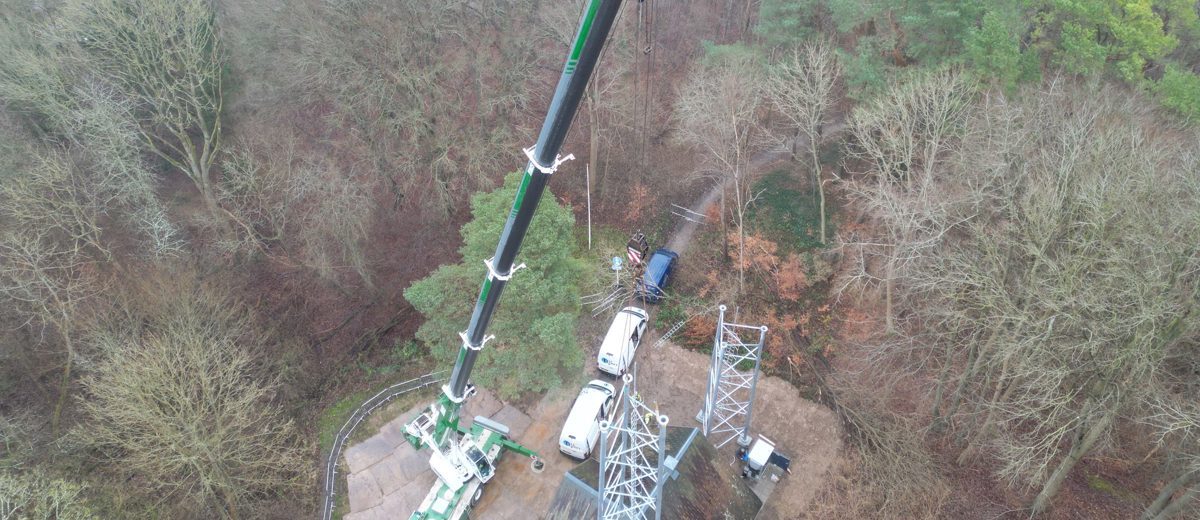 BMS-mobilkran med langt udlæg arbejder ved en telemast omgivet af skov, set fra luften