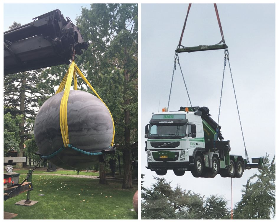 Lastbilkran løfter en stor kunstkugle i en park, mens en anden lastbilkran transporterer et køretøj