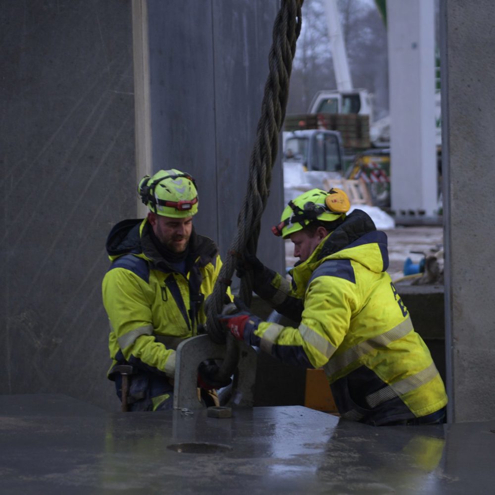 To BMS-riggere fastgør en løftewire til et betonelement på en byggeplads