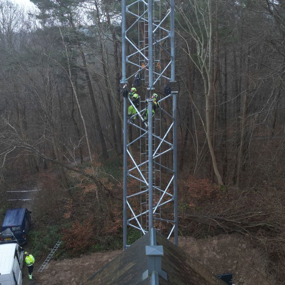 Teknikere arbejder højt oppe på telemast omgivet af skov